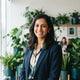 A professional portrait of a smiling South Asian woman in a bright, modern office setting with greenery.